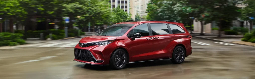 The image shows a red Toyota Sienna minivan driving through an urban street.